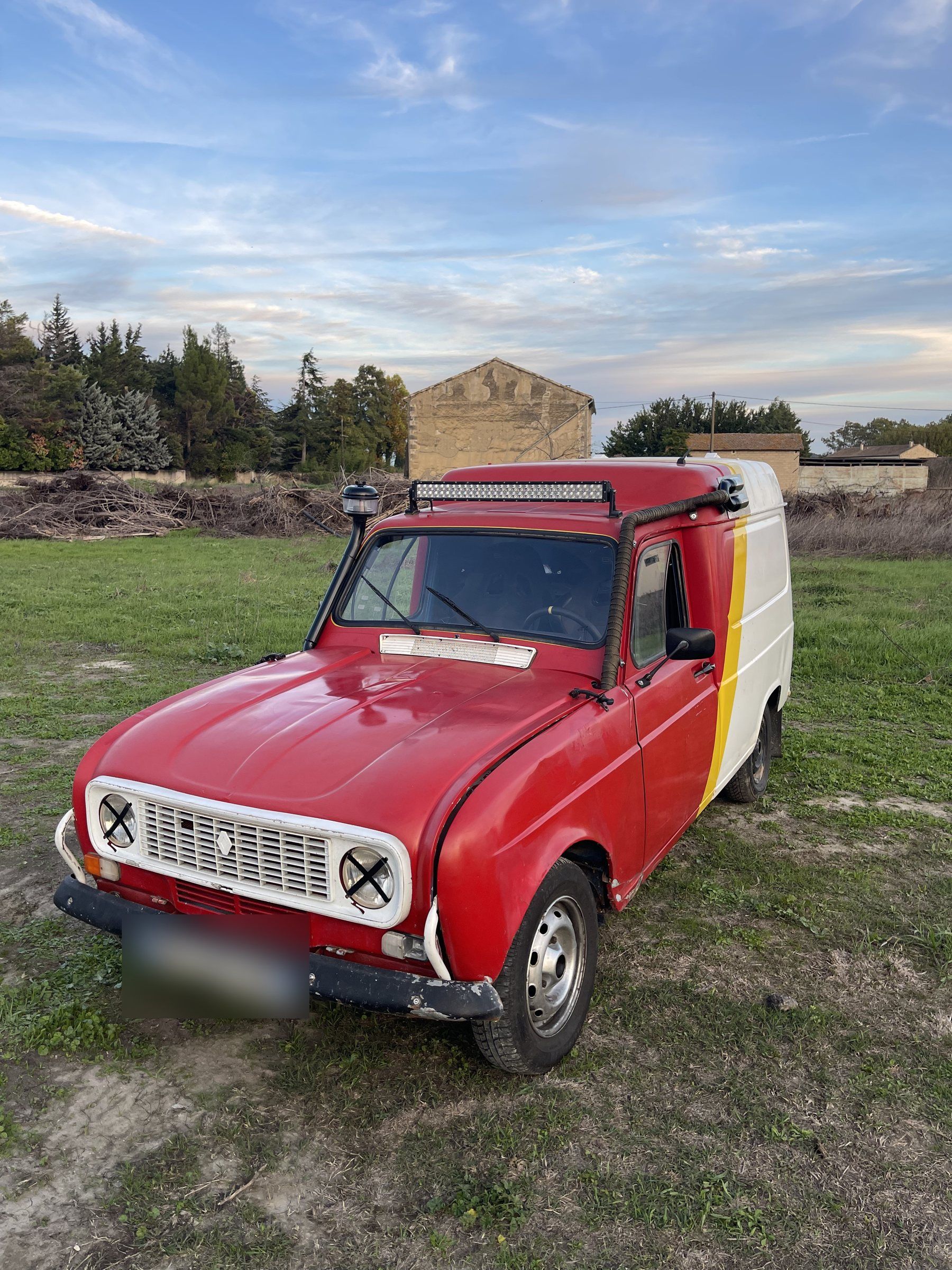 Notre Renault 4L rouge et blanche, rampe de phares sur le toit et snorkel, dans un champ au coucher du soleil.